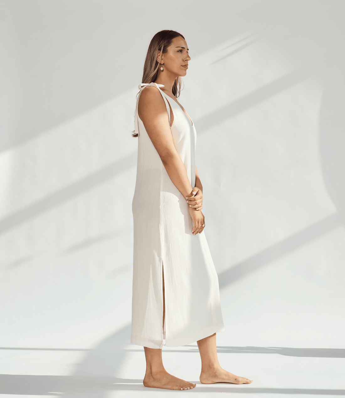 Side view of a woman in a soft cotton dress with tied straps and leg slits against a light background.