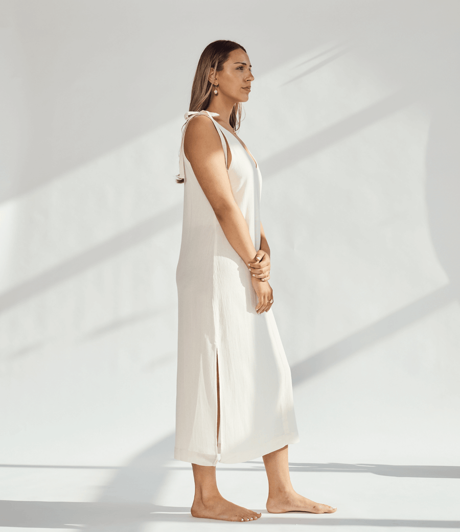 Side view of a woman in a soft cotton dress with tied straps and leg slits against a light background.